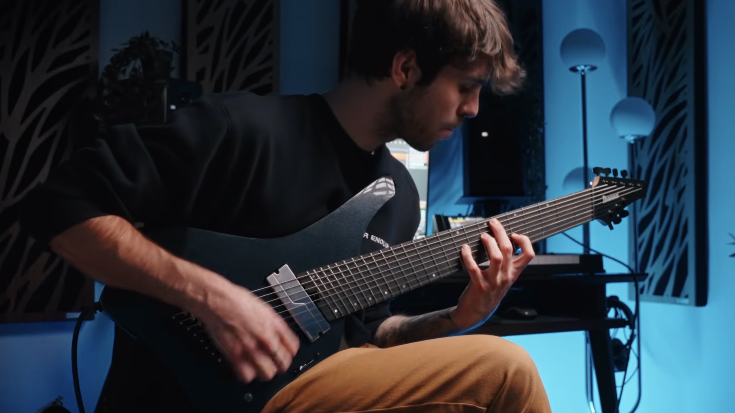 Musician playing an Ibanez Alpha extended-range electric guitar with headstock in a blue-lit studio