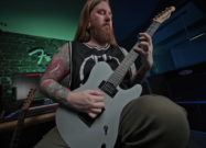 Guitarist with long hair and tattoos playing a matte gray electric guitar in a home studio with colorful lighting and recording equipment.