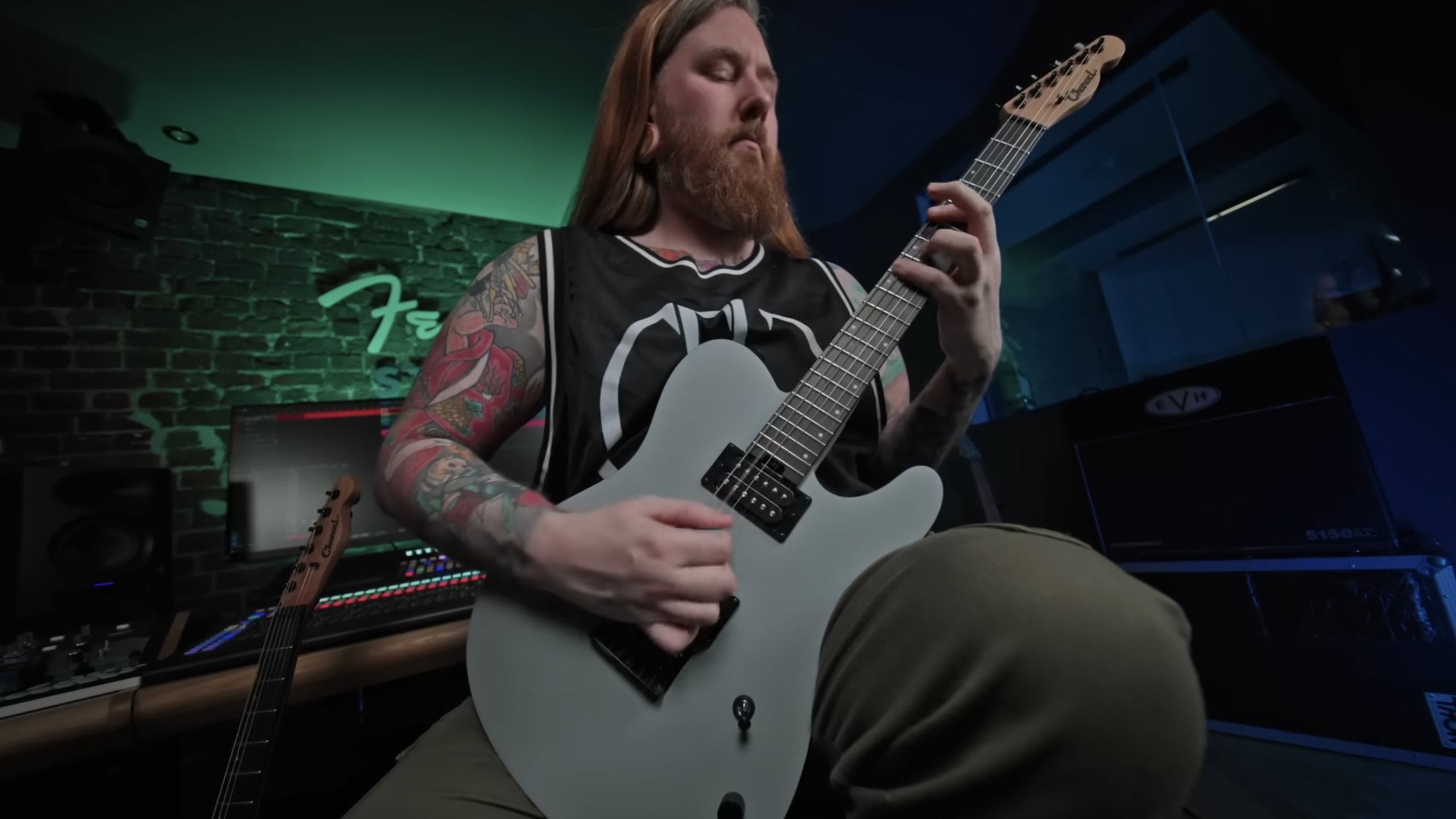 Guitarist with long hair and tattoos playing a matte gray electric guitar in a home studio with colorful lighting and recording equipment.