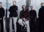 Erra band members posing in a softly lit industrial room with sheer curtains and natural light.