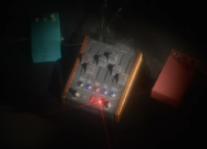 Studio photo of a boutique stereo delay guitar pedal with wooden sides and multiple sliders, glowing LED indicators, and connected cables, surrounded by two smaller effects pedals in teal and red on a dark background with dramatic lighting.