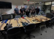 Group of men standing behind a conference table filled with multiple recovered bass guitars in a meeting room, following a theft recovery in Las Vegas.