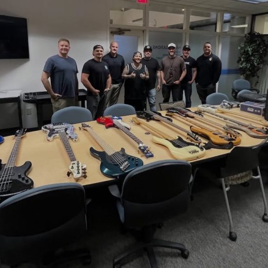Group of men standing behind a conference table filled with multiple recovered bass guitars in a meeting room, following a theft recovery in Las Vegas.