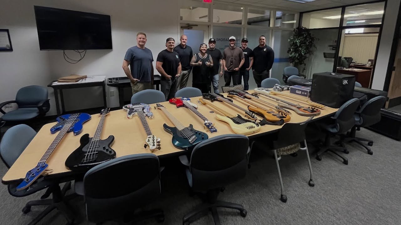 Group of men standing behind a conference table filled with multiple recovered bass guitars in a meeting room, following a theft recovery in Las Vegas.