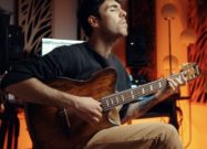 Musician playing an Ibanez FRH20 nylon-string electric-acoustic guitar in a warmly lit recording studio with studio monitors and ambient orange lighting in the background.