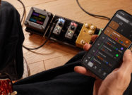 Person holding a smartphone running the TONEX Control app while sitting with an electric guitar and a pedalboard featuring TONEX ONE+ units on the floor.