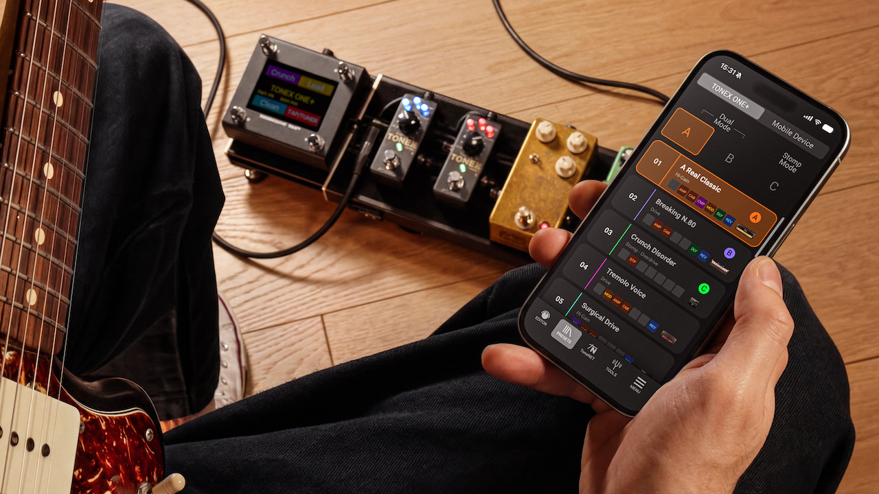 Person holding a smartphone running the TONEX Control app while sitting with an electric guitar and a pedalboard featuring TONEX ONE+ units on the floor.