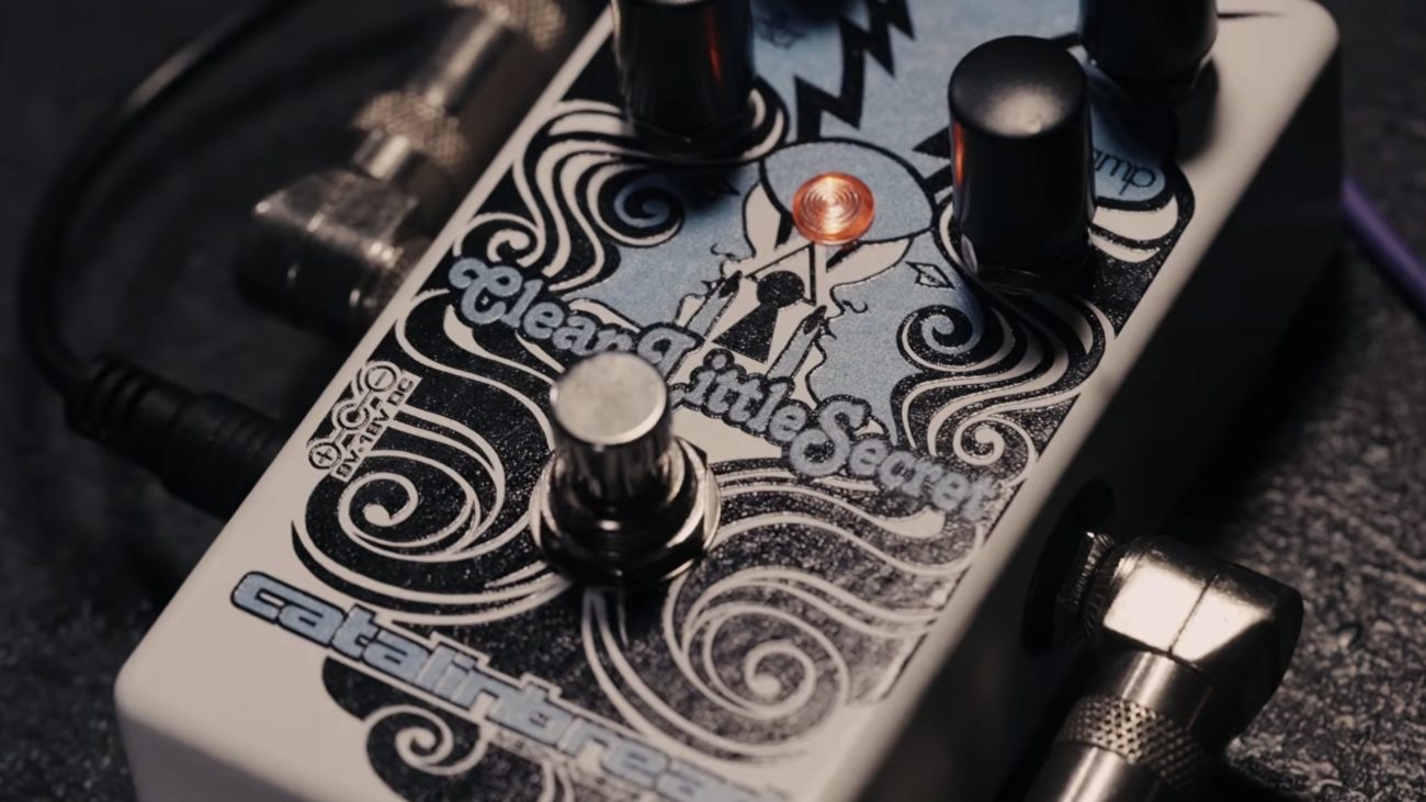 Close-up of a Catalinbread Clean Little Secret guitar effects pedal with decorative artwork, glowing LED indicator, footswitch, and control knobs, connected with instrument cables.