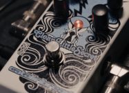 Close-up of a Catalinbread Clean Little Secret guitar effects pedal with decorative artwork, glowing LED indicator, footswitch, and control knobs, connected with instrument cables.