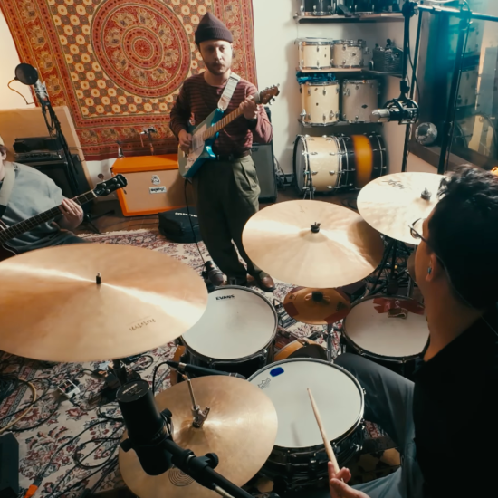 Three musicians perform an instrumental progressive rock cover of Radiohead’s “Everything In Its Right Place” inside a recording studio, featuring guitar, bass, and drums surrounded by recording equipment and drum kits.