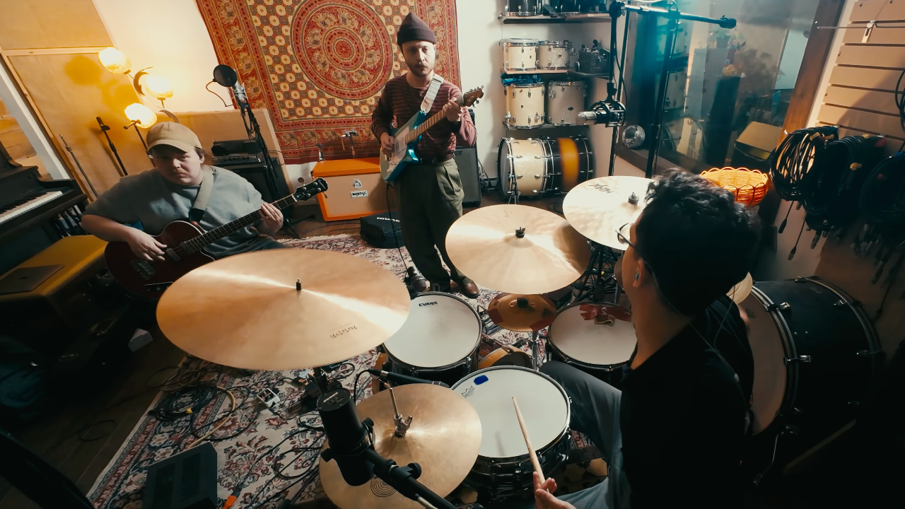Three musicians perform an instrumental progressive rock cover of Radiohead’s “Everything In Its Right Place” inside a recording studio, featuring guitar, bass, and drums surrounded by recording equipment and drum kits.