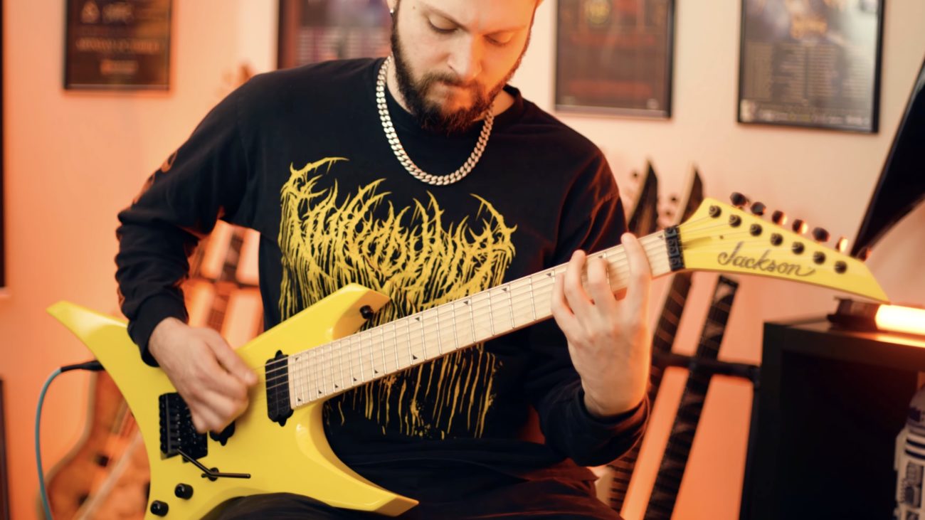 Sam Mooradian playing a bright yellow Jackson extended-range electric guitar in a warm-lit home studio, wearing a black metal band long sleeve shirt and silver chain necklace.