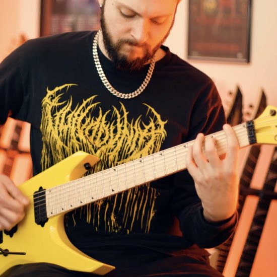 Sam Mooradian playing a bright yellow Jackson extended-range electric guitar in a warm-lit home studio, wearing a black metal band long sleeve shirt and silver chain necklace.
