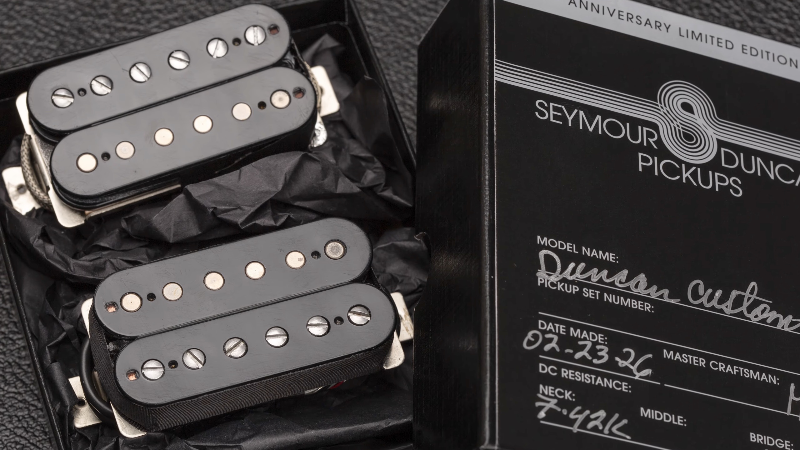 Close-up of a limited-edition Seymour Duncan 50th Anniversary Duncan Custom pickup set resting in black packaging beside the commemorative box. The image shows two humbucker pickups with black bobbins, exposed pole pieces, and vintage-style detailing, highlighting the hand-built Custom Shop presentation.